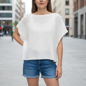 Könnte beinhalten: Ein weißes Oberteil aus offen gewebtem Strick mit Bootsausschnitt und kurzen Ärmeln. Dazu werden blaue Jeansshorts getragen. Das Outfit ist lässig und für warmes Wetter geeignet. Im Hintergrund ist eine Stadtstraße zu sehen.