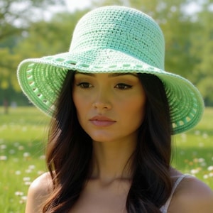 May include: A light green crochet sun hat with a wide brim and a decorative edge. The hat is worn by a person with long, dark hair. The background is a blurred outdoor scene with trees and grass.