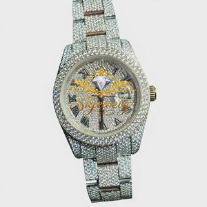 May include: A silver-toned wristwatch fully encrusted with small clear stones on the bezel, case, and link bracelet. The watch face features Roman numeral markers, a central gold-colored emblem with a diamond graphic, and the text Skyjewely.
