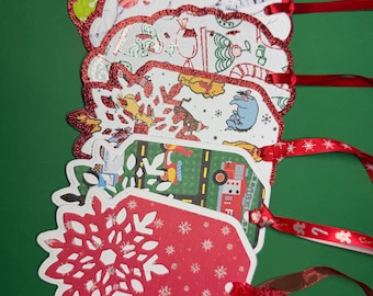 May include: A stack of Christmas gift tags with red ribbon. The tags have scalloped edges and feature festive patterns, including snowflakes, cartoon characters, and holiday scenes. The tags are on a green background.