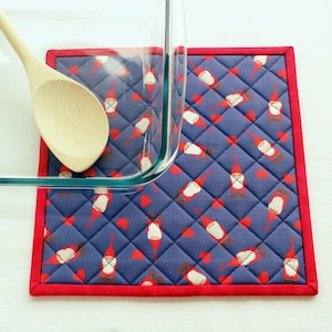 May include: A blue quilted hot pad with a red border. The pad is decorated with a pattern of red and white gnomes and red hearts. A glass baking dish and wooden spoon are on top of the hot pad.