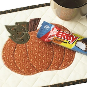 Peut inclure: Un dessous de verre matelassé en forme de citrouille avec un motif en tissu brun, vert et orange. Une barre chocolatée avec le texte "Derby" et "Lait & Noix de coco" est posée sur le dessous de verre.