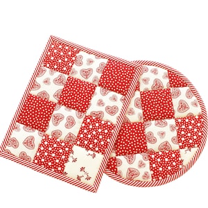 May include: A quilted potholder and trivet set. The set features a patchwork design with red and white squares. Some squares have red polka dots, others have red hearts, and some have a floral pattern. The edges are trimmed with red and white striped fabric.