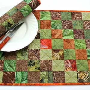 Könnte beinhalten: Ein grünes und braunes Patchwork-Platzset mit orangefarbenem Rand. Das Platzset besteht aus Stoffquadraten mit verschiedenen Mustern, darunter Blätter und Wirbel. Ein weißer Teller mit Gabel und Messer liegt auf dem Platzset.