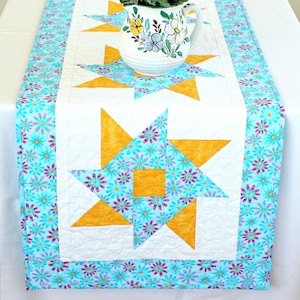 May include: A white table runner with a blue and yellow floral pattern. The runner features a repeating star design with a yellow centre and blue and pink floral accents. The runner has a blue and pink floral border.