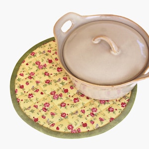 May include: A round quilted pot holder with a green border and a floral pattern of red roses on a yellow background. A beige ceramic pot with a lid is sitting on top of the pot holder.