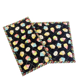 May include: Two quilted potholders featuring a black background adorned with colourful cupcakes and donuts. The edges are trimmed with a striped border in red, yellow, blue, and green. The potholders are square-shaped.
