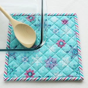May include: A blue quilted hot pad with a snowflake pattern and a white, pink and green striped border. A wooden spoon and a glass baking dish are on top of the hot pad.