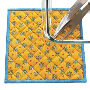 May include: A yellow quilted hot pad with a blue border and a floral pattern. A glass baking dish and two silver utensils are on top of the hot pad.