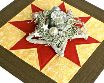 Könnte beinhalten: Ein sternförmiger Tischläufer in Rot und Gold mit braunem Rand. In der Mitte des Läufers befindet sich ein dekoratives Mittelstück mit Zweigen, Grün, Tannenzapfen und silbernen Ornamenten.