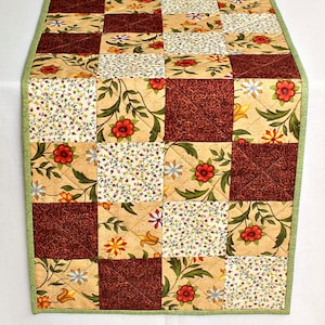 May include: A patchwork table runner with a green border. The runner is made of various fabrics with floral patterns in shades of brown, yellow, and white.