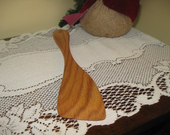May include: A wooden spatula with a light brown color and a slightly curved shape. The spatula is lying on a white lace tablecloth.