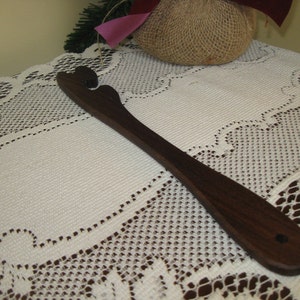 May include: A wooden kitchen utensil with a unique shape, featuring a curved handle and a flat, rounded head with a small hole. The utensil is made of dark brown wood and has a smooth finish.