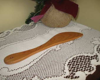 May include: A wooden spatula with a rounded edge and a hole in the handle. The spatula is light brown and is lying on a white lace tablecloth.