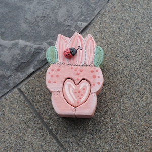 May include: A pink wooden flower pot with a heart-shaped opening and a ladybug on top. The pot has a green leaf design and white polka dots.