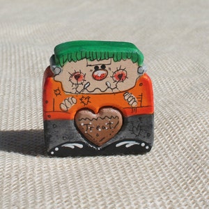 May include: A hand-painted wooden figurine of a Frankenstein monster holding a heart-shaped treat that says "Treat". The monster is wearing an orange shirt, black pants, and has green hair.