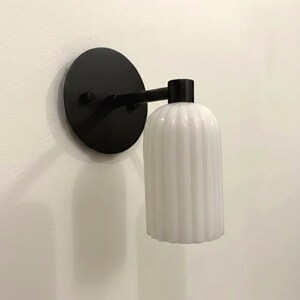 May include: A wall-mounted light fixture featuring a circular matte black base and a horizontal arm extending to a vertical, cylindrical white glass shade with a distinct vertical ribbed texture throughout its surface.