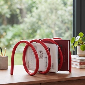 May include: A bright red metal book rack featuring a continuous spiral or coil design. The structure consists of three rounded loops that hold two books upright. The rack is positioned on a wooden surface, showcasing a glossy finish and smooth curves.