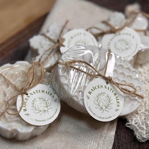 May include: Several handmade soaps are displayed, some wrapped in clear plastic and tied with twine. Each soap has a unique shape and is adorned with a round tag that reads "Raíces Naturales, Naturally Crafted Goods."