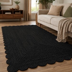 May include: A rectangular black area rug featuring a braided texture and a decorative scalloped edge. The rug is constructed from a woven material and lies flat on a wooden floor, showcasing a uniform dark color throughout its entire surface.