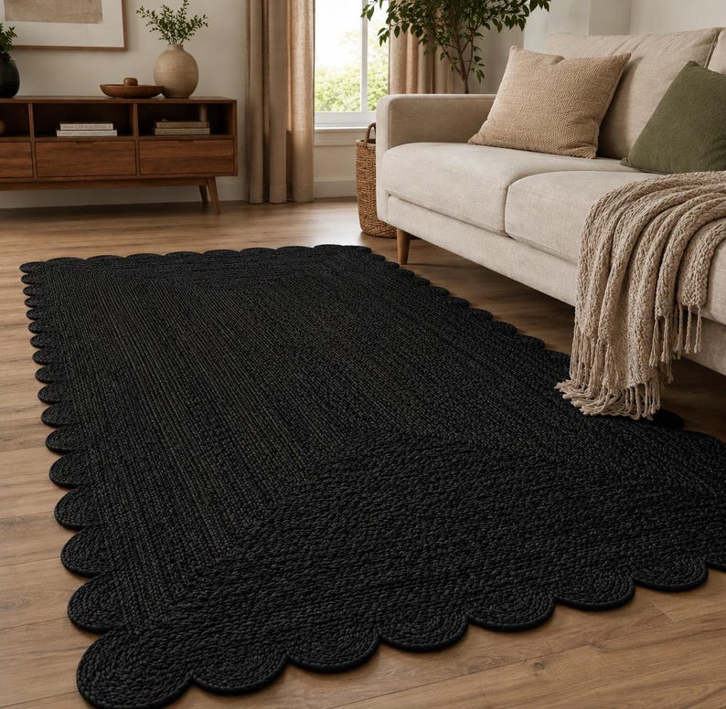 May include: A rectangular black area rug featuring a braided texture and a decorative scalloped edge. The rug is constructed from a woven material and lies flat on a wooden floor, showcasing a uniform dark color throughout its entire surface.