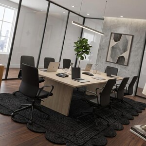 May include: A square light wood conference table surrounded by eight black office chairs on a large black scalloped rug. The table holds several laptops, a conference phone, and a glass carafe. A tall green plant sits in the center of the table.