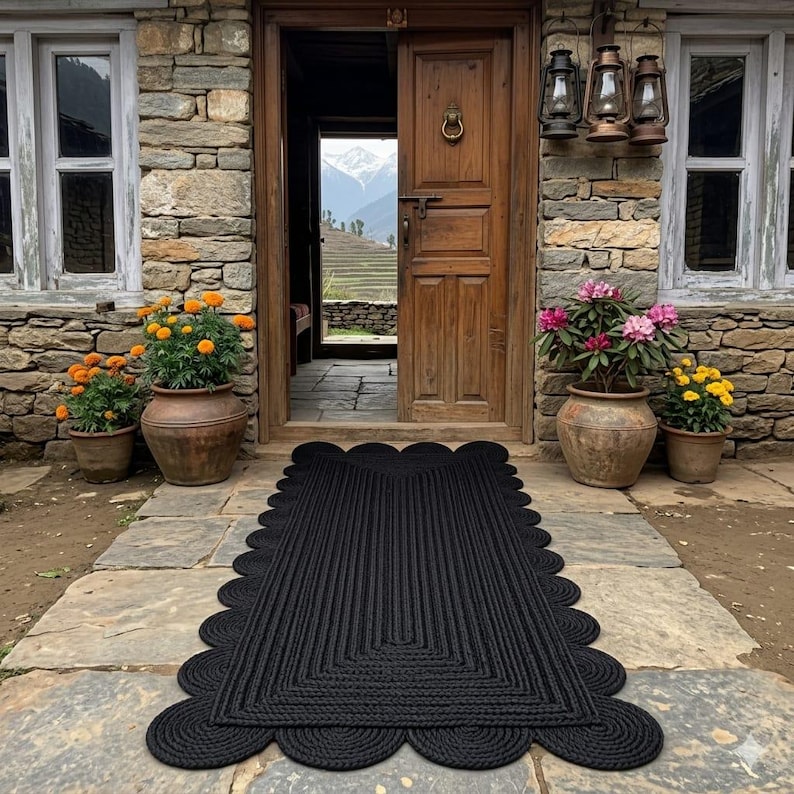 May include: A rectangular black braided rug with a scalloped edge design, positioned on a stone patio in front of a wooden door. The rug features a dense, textured weave pattern that follows the rectangular shape, framed by a series of rounded loops.
