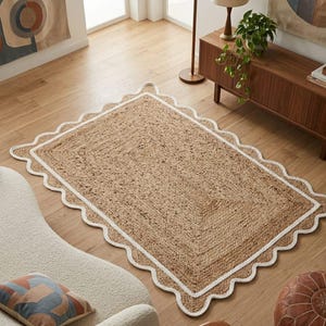 May include: A rectangular area rug made of natural tan woven jute fibers. The rug features a distinct scalloped edge border outlined in a thick, solid white fabric trim that frames the textured, earthy brown center of the floor covering.