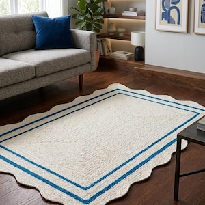 May include: A rectangular, off-white rug with a scalloped edge and two parallel blue stripes. The rug is made of a textured material and is placed on a dark wood floor. The rug is in a living room setting.