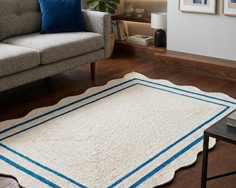 Op de afbeelding: Een rechthoekig, crèmekleurig tapijt met een geschulpte rand en twee blauwe strepen. Het tapijt is gemaakt van een textuurrijk materiaal en ligt op een donkere houten vloer. Het tapijt bevindt zich in een woonkamer.