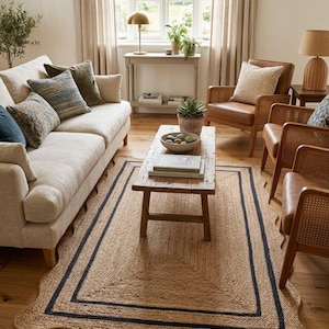 May include: A living room scene featuring a natural jute rug with a scalloped edge and a dark blue border. The room includes a beige sofa with decorative pillows, leather armchairs, and a wooden coffee table. A gold lamp sits on a side table.