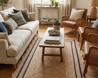 Op de afbeelding: Woonkamer met een natuurlijk jute vloerkleed met een geschulpte rand en een donkerblauwe rand. De kamer omvat een beige bank met sierkussens, leren fauteuils en een houten salontafel. Een gouden lamp staat op een bijzettafel.