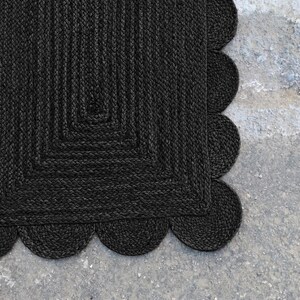 May include: A rectangular rug crafted from dark charcoal black braided fibers. The rug features a distinct central rectangular pattern composed of concentric braided lines, finished with a decorative scalloped edge border made of circular braided loops.