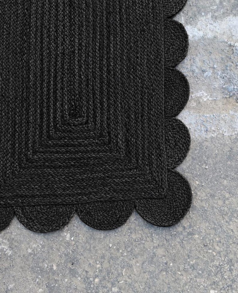 May include: A rectangular rug crafted from dark charcoal black braided fibers. The rug features a distinct central rectangular pattern composed of concentric braided lines, finished with a decorative scalloped edge border made of circular braided loops.
