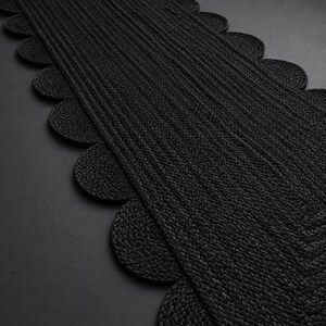 May include: A long, rectangular black table runner featuring a textured, braided construction. The edges of the runner are finished with a series of repeating, rounded scalloped shapes that extend along both sides of the central woven panel.