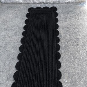 May include: A rectangular black runner rug featuring a braided texture and a scalloped edge design. The rug is constructed from concentric rows of dark material that follow the rectangular shape, finished with a series of circular loops along the sides.