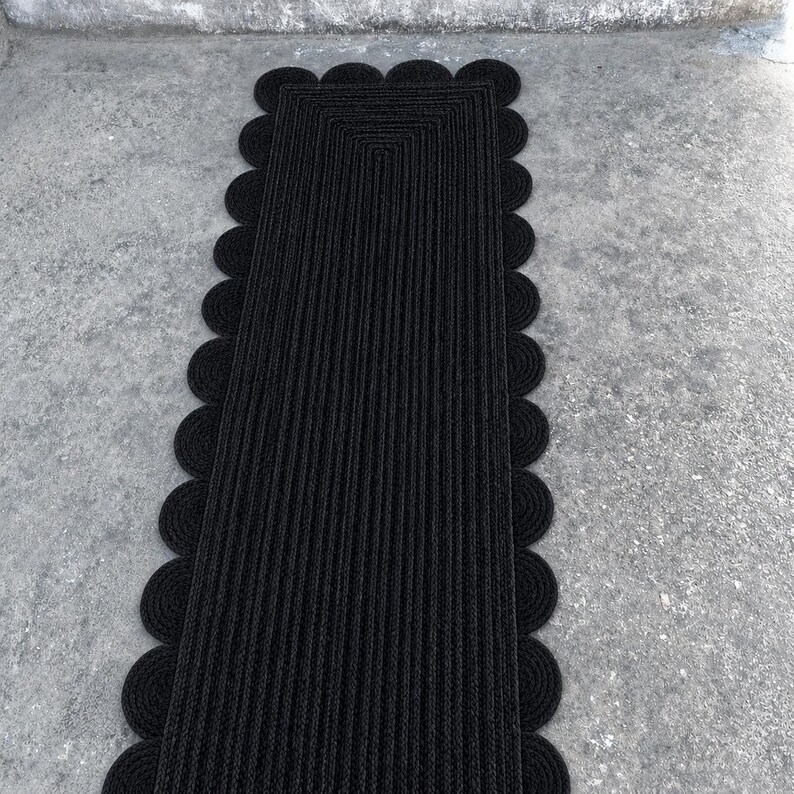 May include: A rectangular black runner rug featuring a braided texture and a scalloped edge design. The rug is constructed from concentric rows of dark material that follow the rectangular shape, finished with a series of circular loops along the sides.
