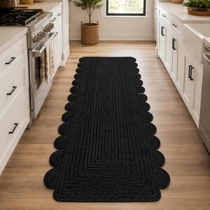 May include: A long, rectangular runner rug in solid black, featuring a braided texture and a decorative scalloped edge design that runs along both sides of the length. The rug is positioned centrally on a light-colored wooden floor in a kitchen.