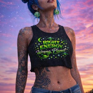 Puede incluir: Un top corto negro con el texto "RIGHT ENERGY WRONG PLANET" en verde y blanco, combinado con vaqueros azules. La imagen presenta a una persona con cabello azul y purpurina, sobre un cielo colorido.