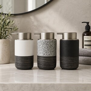 Può includere: Tre dispenser di sapone in ceramica con pompe in acciaio inossidabile. I dispenser sono bianchi, grigio maculato e neri, ciascuno con una base scanalata grigio scuro. I dispenser sono disposti su un piano di lavoro in marmo.