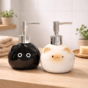 May include: Two ceramic soap dispensers. One is black with white eyes, the other is white with tan ears and a winking eye. Both have silver pump tops and are on a wooden surface. Bathroom decor.