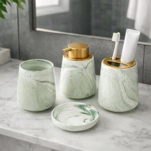 Puede incluir: Un juego de baño de cuatro piezas con diseño de mármol. El juego incluye un vaso, un dispensador de jabón con bomba dorada, un soporte para cepillos de dientes y un jabonera pequeña. Las piezas son blancas con vetas verdes y detalles dorados.
