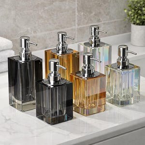 Può includere: Sei dispenser di sapone decorativi in vetro con erogatori cromati. I dispenser sono quadrati e sono disponibili in vari colori, tra cui nero, ambra e iridescente. Sono disposti su un piano di lavoro bianco.
