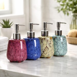 Può includere: Quattro dispenser di sapone in ceramica rosa, blu, giallo e verde acqua. Ogni dispenser ha un motivo maculato e una pompa argentata. I dispenser sono disposti in fila su un piano di lavoro in marmo.