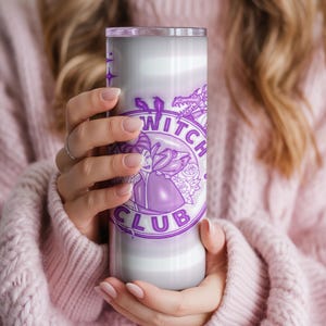May include: A tall, cylindrical tumbler with a purple and white design. The tumbler features the words "Witch Club" in a circular design, along with a witch illustration and a dragon. The tumbler is held by a person wearing a pink sweater.