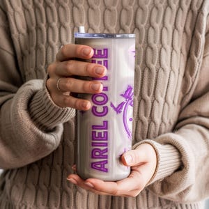 May include: A stainless steel tumbler with a purple design and the text "ARIEL COFFEE". The tumbler has a white straw and a silver lid. The design includes a clock and birds.