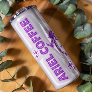 May include: A white and purple insulated tumbler with the words "ARIEL COFFEE" in a purple, stylized font. The tumbler features a design of a mermaid and stars. The tumbler is cylindrical and has a silver lid.