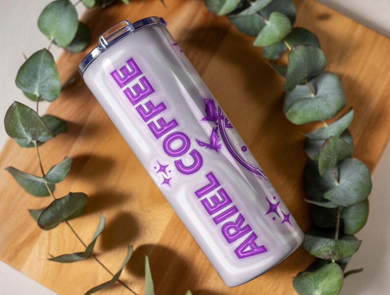 May include: A white and purple insulated tumbler with the words "ARIEL COFFEE" in a purple, stylized font. The tumbler features a design of a mermaid and stars. The tumbler is cylindrical and has a silver lid.