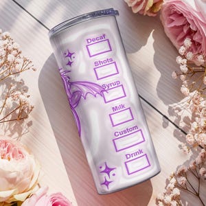 May include: A white tumbler with a purple dragon design and text boxes for drink customization. The tumbler includes labels for Decaf, Shots, Syrup, Milk, Custom, and Drink. The background features pink roses and white flowers.