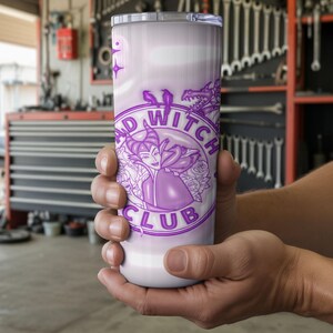 May include: A white and lavender tumbler featuring the text "BAD WITCH CLUB" in a circular design with a purple illustration of a witch. The tumbler is held in a person's hands. The background is a garage.